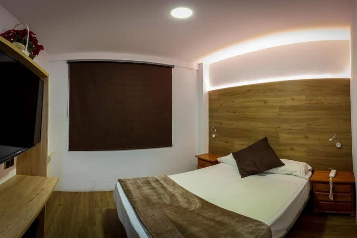 Ein Schlafzimmer mit einem hölzernen Kopfteil, einem Bett mit weißer Matratze und brauner Decke, einem Nachttisch mit Lampe sowie einem an der Wand befestigten Fernseher.