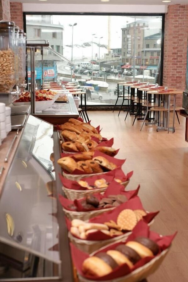 A buffet with various food items, a large window with a view of buildings and parked cars, and a brick wall. - Hotel Les Neus (Pas de la Casa)