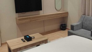 A hotel room with a bed, a television mounted on the wall, a small table with a remote control, a gray armchair, and a mirror. - Rama Dubai Serviced Apartments 4 (Riyadh)