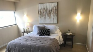 Un lit bien fait, recouvert d’une couette grise, deux tables de chevet avec des lampes, un tableau abstrait accroché au mur et une fenêtre avec des stores.