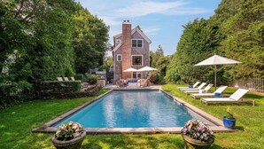 A backyard with a swimming pool, a brick house, and a patio with lounge chairs and umbrellas. - Elegant Coastal Escape with Expansive Yard, Heated Pool, and Outdoor... (Water Mill)