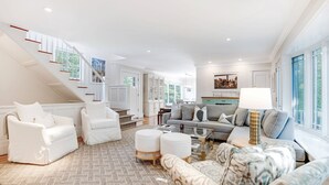 A spacious living room with a staircase, a grey sofa, white armchairs, and a coffee table. - Elegant Coastal Escape with Expansive Yard, Heated Pool, and Outdoor... (Water Mill)