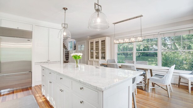 A modern kitchen with a large island, white cabinetry, stainless steel appliances, and a dining area with wicker chairs and a view of the outdoors. - Elegant Coastal Escape with Expansive Yard, Heated Pool, and Outdoor... (Water Mill)