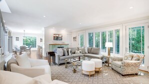 A spacious living room with a large sofa, a coffee table, and a patterned armchair. There is a dining area with wicker chairs and a table, and a kitchen area with a green cabinet. - Elegant Coastal Escape with Expansive Yard, Heated Pool, and Outdoor... (Water Mill)