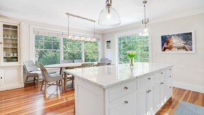 A spacious kitchen with a large island, white cabinetry, and a view of greenery through large windows. - Elegant Coastal Escape with Expansive Yard, Heated Pool, and Outdoor... (Water Mill)