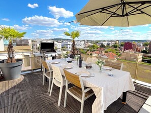 A rooftop terrace with a table set for dining, a grill, and potted plants.