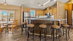 A kitchen with wooden cabinets, a stainless steel refrigerator, a large island with stools, and a dining area with a table and chairs. - Navigate Stays Pet Friendly Desert Dream (Yucca Valley)