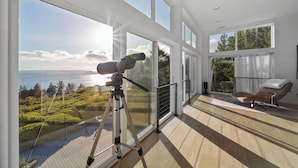 A balcony with a telescope, large windows, a chair, and a view of the sea. - Port Orford Ocean View, Waterfront & BBQ Area (Port Orford)