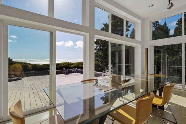 A modern dining area with a glass table and chairs, large windows offering views of a deck and trees.