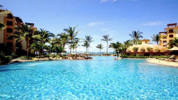 A large, clear swimming pool surrounded by palm trees and hotel buildings.