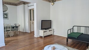 A living room with a TV on a white stand, a dining area with a table and chairs, and a sofa with a green throw.