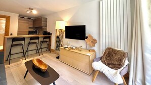 A compact hotel room with a kitchenette, a flat-screen TV, a wooden sideboard, and a cozy seating area with a fur throw.