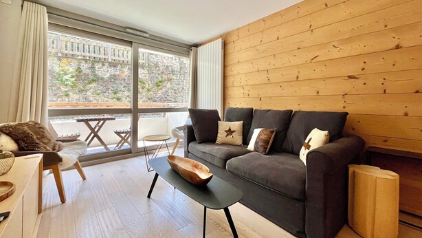 A modern living room with a dark grey sofa, wooden floor, and a large window offering a view of a stone wall and greenery outside.