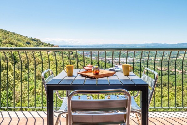 A balcony with a table and chairs, a tray with a teapot and cups, and a scenic view of greenery and distant mountains.