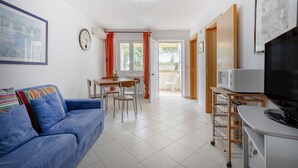 A living room with a sofa, a dining table, chairs, a TV, and a kitchen area with a microwave. - GuestHost - Villa Belvedere: Apartment E (Ostuni)