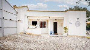 A building with a stone-tiled courtyard, a garage door, and a small entrance with a bench and potted plant. - GuestHost - Villa Belvedere: Apartment E (Ostuni)