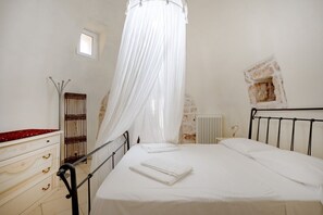 A bedroom with a bed, white curtains, a radiator, a small window, and a dresser with drawers.