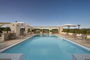 Une piscine rectangulaire à l’eau d’un bleu limpide, entourée de chaises longues et de parasols, avec une terrasse en pierre et en béton.