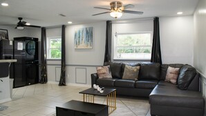 A modern living room with a sectional sofa, a coffee table, a ceiling fan, and a refrigerator. - Modern In-Law Suite Near Jax Beaches & Mayo (Jacksonville)