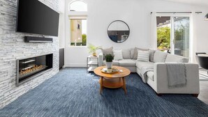 A modern living room with a flat-screen TV mounted on a white brick wall, a fireplace, a round coffee table, a white sofa, and a large window offering a view of greenery. - Private Hilltop Escape: Pool, Hot Tub, Near Beach (Vista)