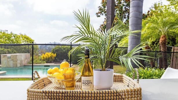 A wicker tray with a bottle of California Roots Chardonnay, a bowl of lemons, and glasses on a table with a view of a pool and greenery. - Private Hilltop Escape: Pool, Hot Tub, Near Beach (Vista)