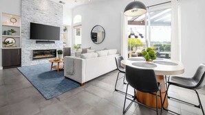 A modern living room with a fireplace, a flat-screen TV mounted on a stone wall, a round dining table with chairs, and a large window with a view of greenery. - Private Hilltop Escape: Pool, Hot Tub, Near Beach (Vista)
