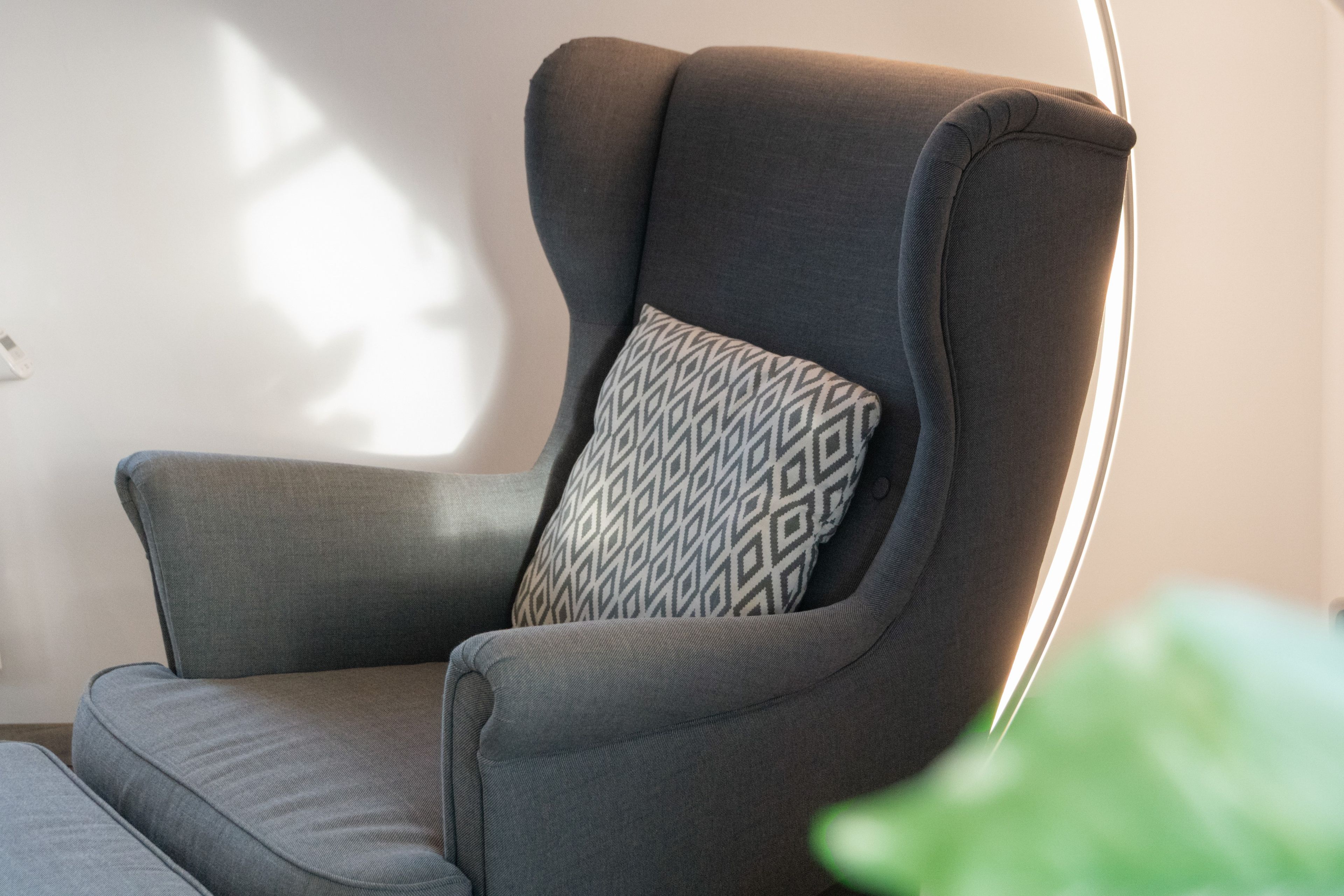 Un sillón tapizado color gris con un cojín estampado, ubicado frente a una pared donde una fuente de luz proyecta una sombra.
