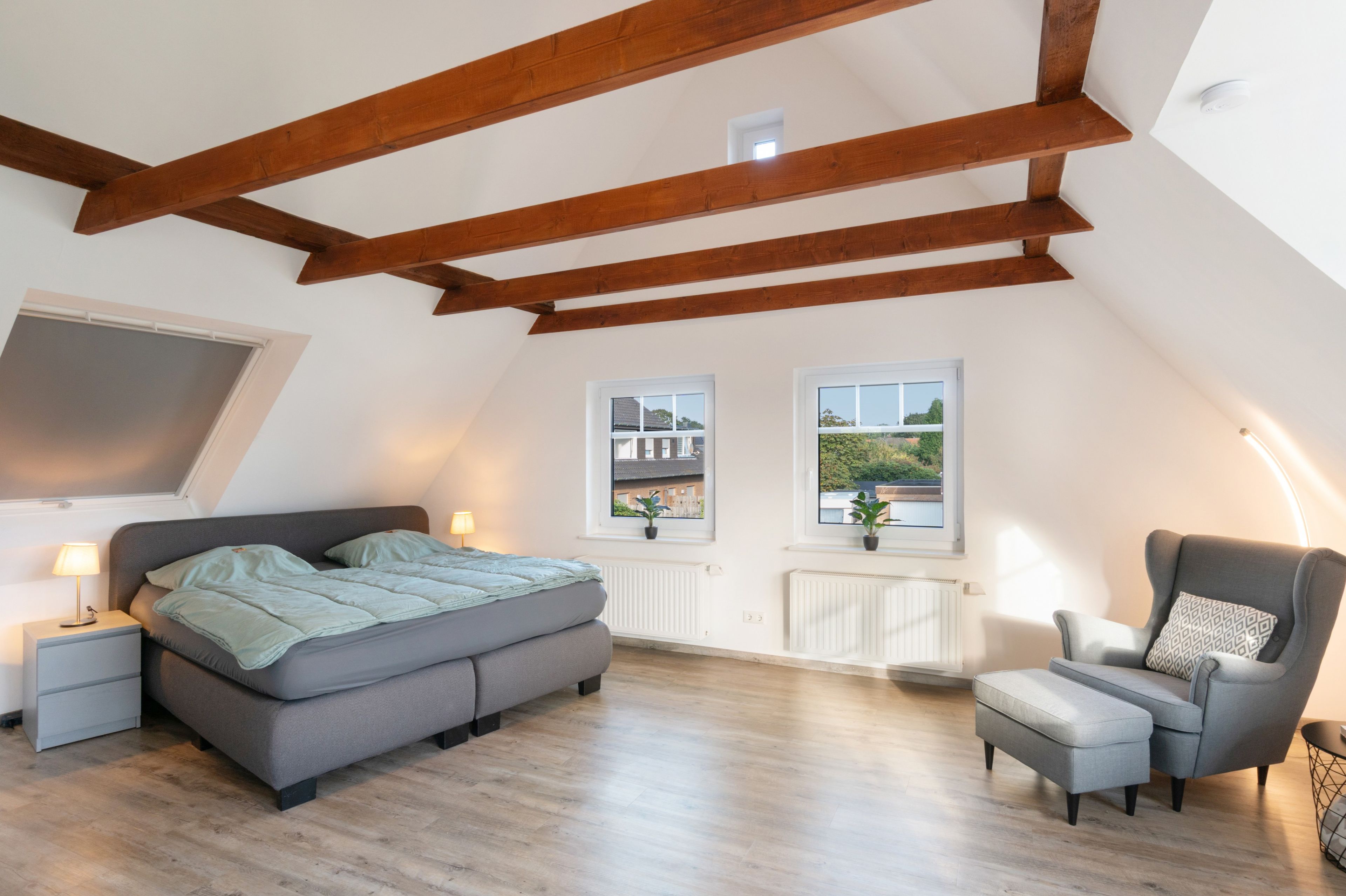 Un dormitorio con una cama, mesitas de noche, una silla y dos ventanas.