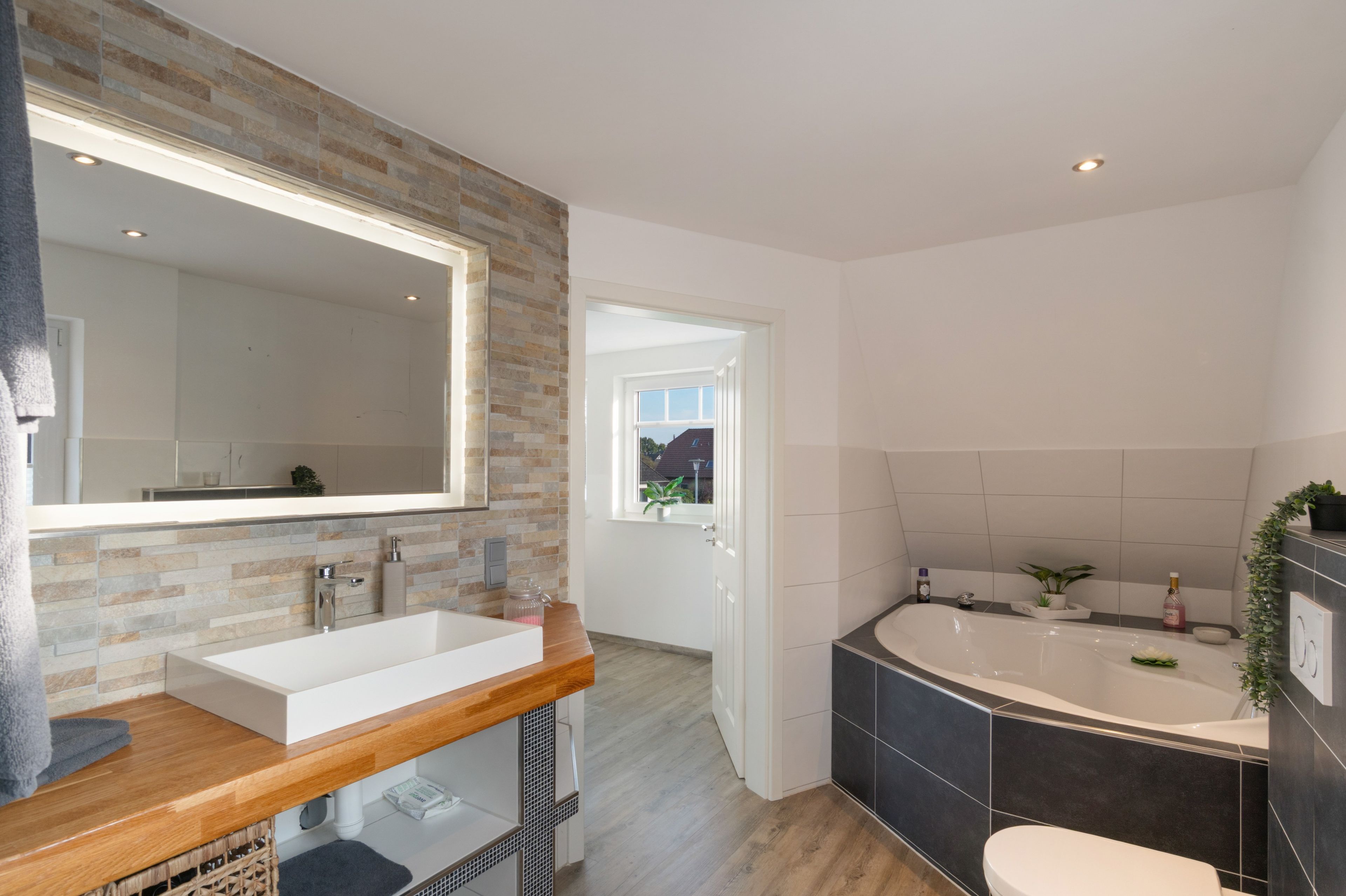 Ein modernes Badezimmer mit einem großen Spiegel, einer Holzwaschmaschine, einer freistehenden Badewanne und Blick ins Freie.