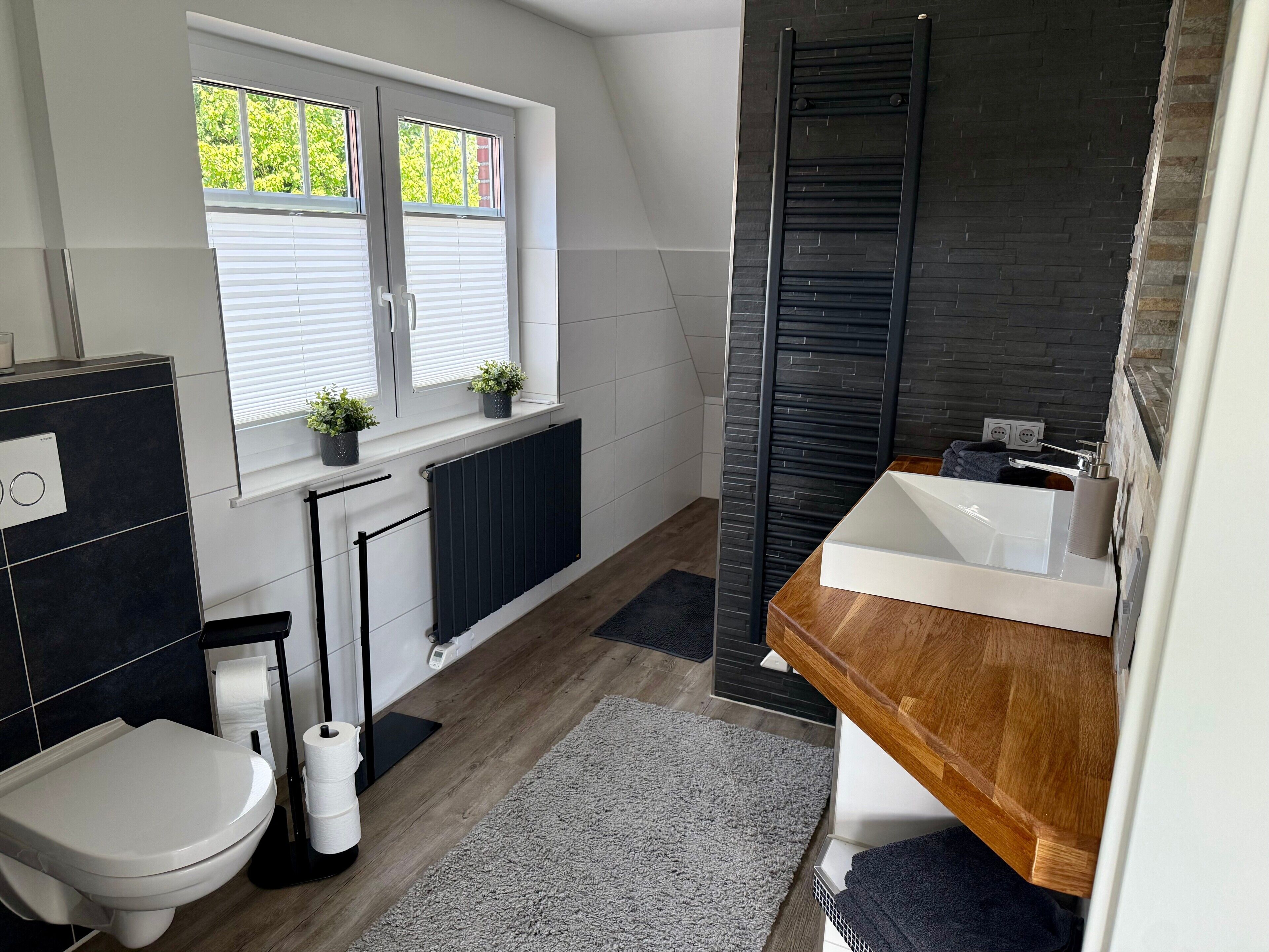Een moderne badkamer met een houten wastafelmeubel, een wit toilet, een zwart bad en een raam met zonwering.