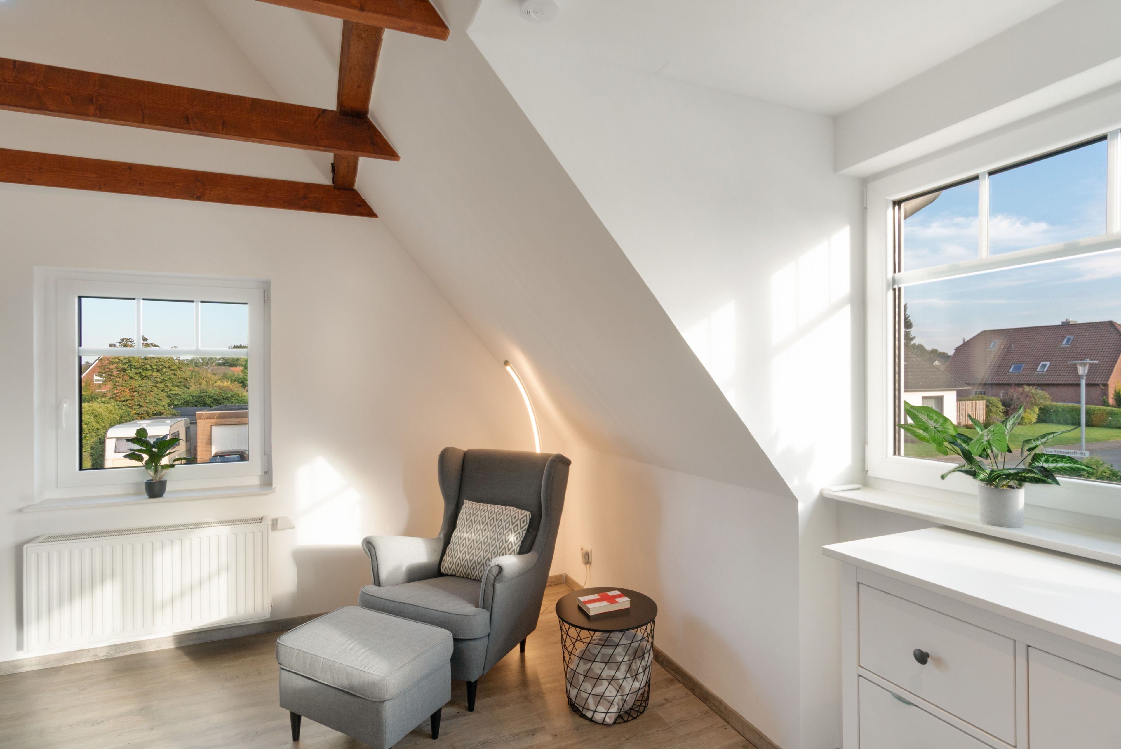 A modern loft bedroom with a sloped ceiling, a grey sofa, a bedside table with a lamp, and a view of a residential area.