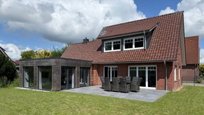 Et moderne hus i to etager med tegltag, store vinduer og en terrasse med udendørs møbler.