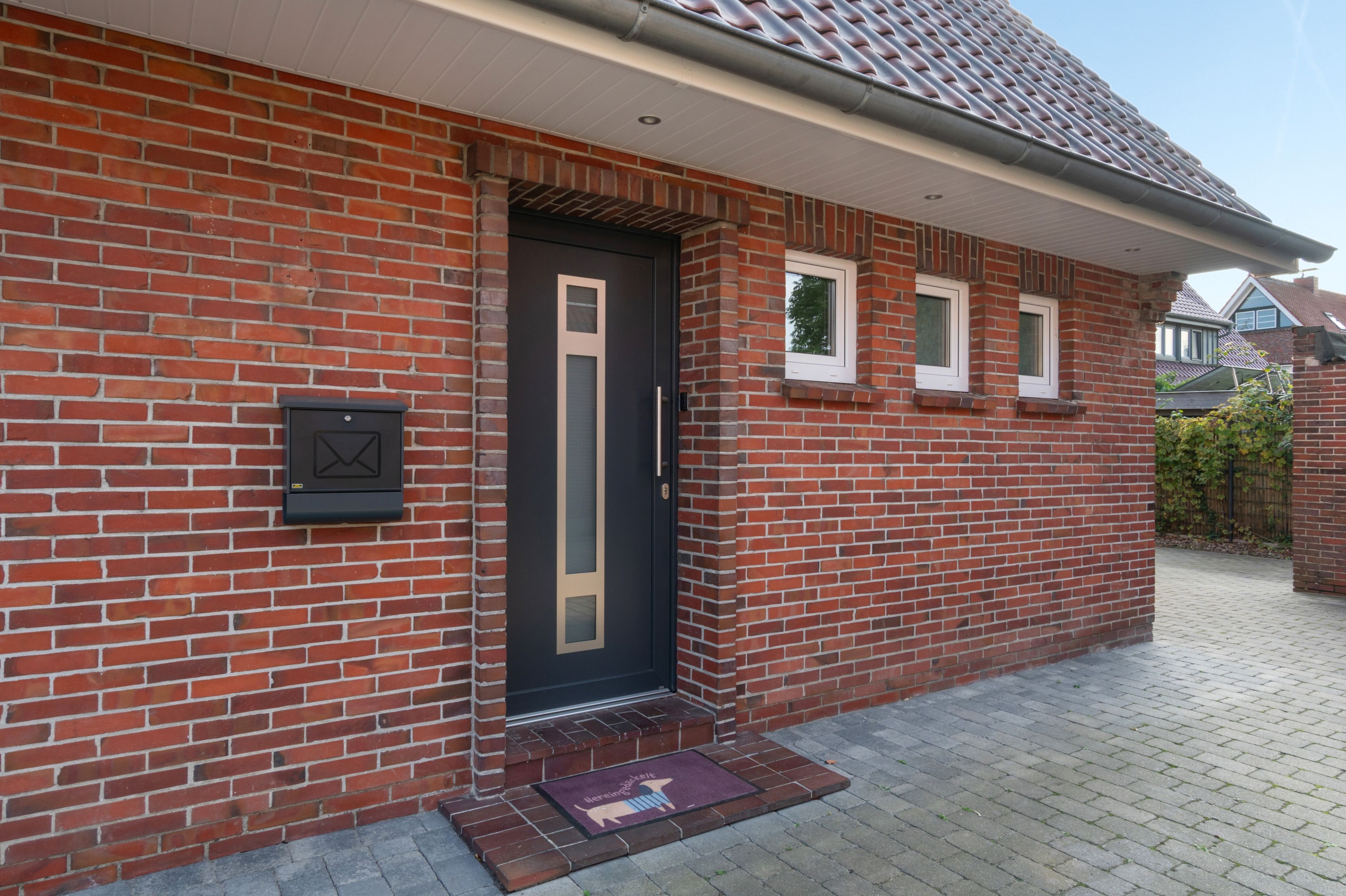 Une maison en briques avec une porte d’entrée sombre, une boîte aux lettres et un petit porche doté d’une fenêtre.
