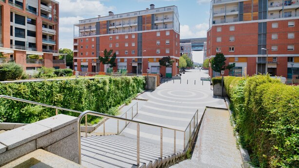 Ein mehrstöckiges Wohngebäude mit Balkonen, einer zentralen Treppe und einer begrünten Anlage mit Hecken.