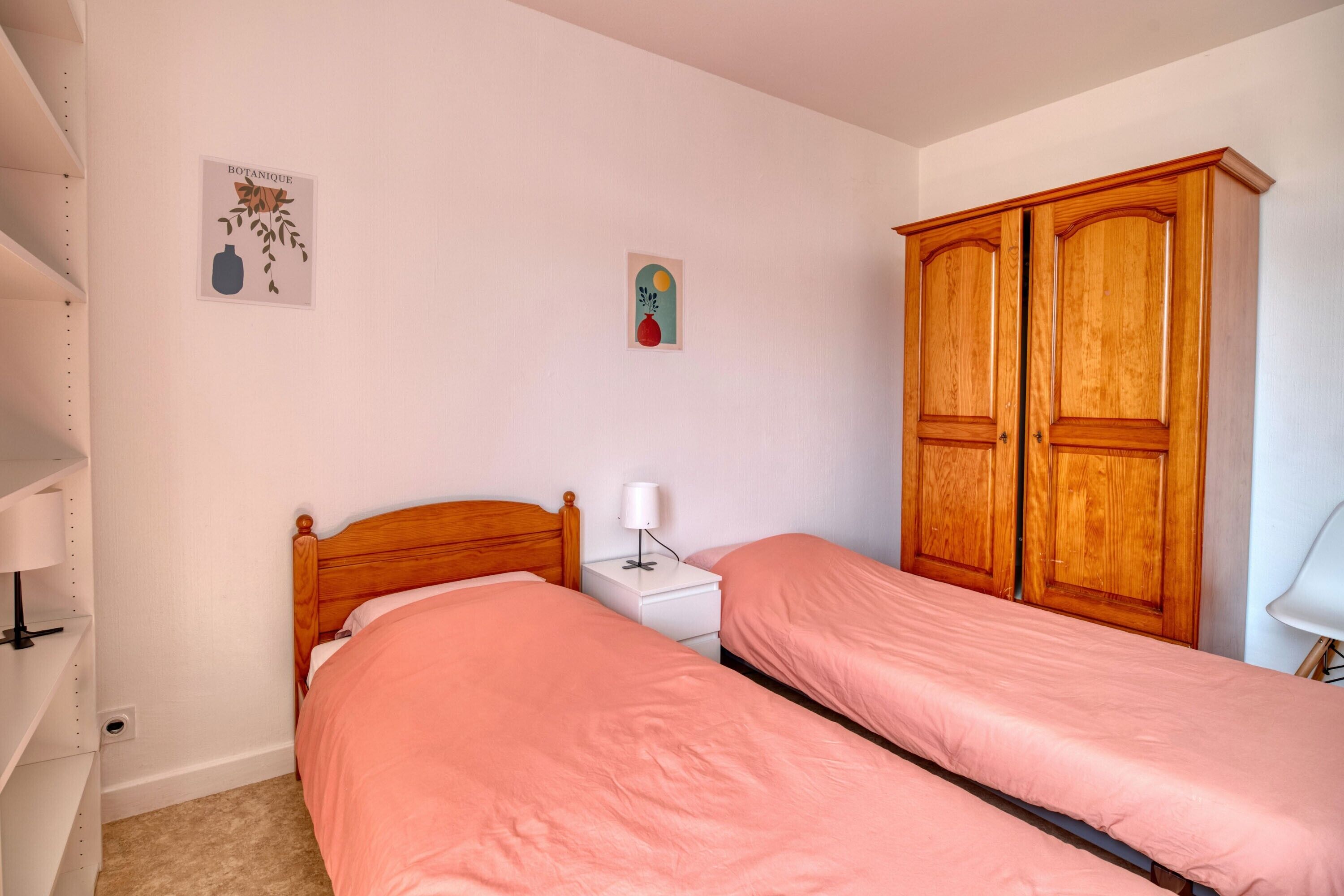 A bedroom with two single beds, a wooden wardrobe, a small bedside table with a lamp, and a wall decoration.