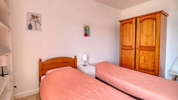 A bedroom with two single beds, a wooden wardrobe, a small bedside table with a lamp, and a wall decoration.
