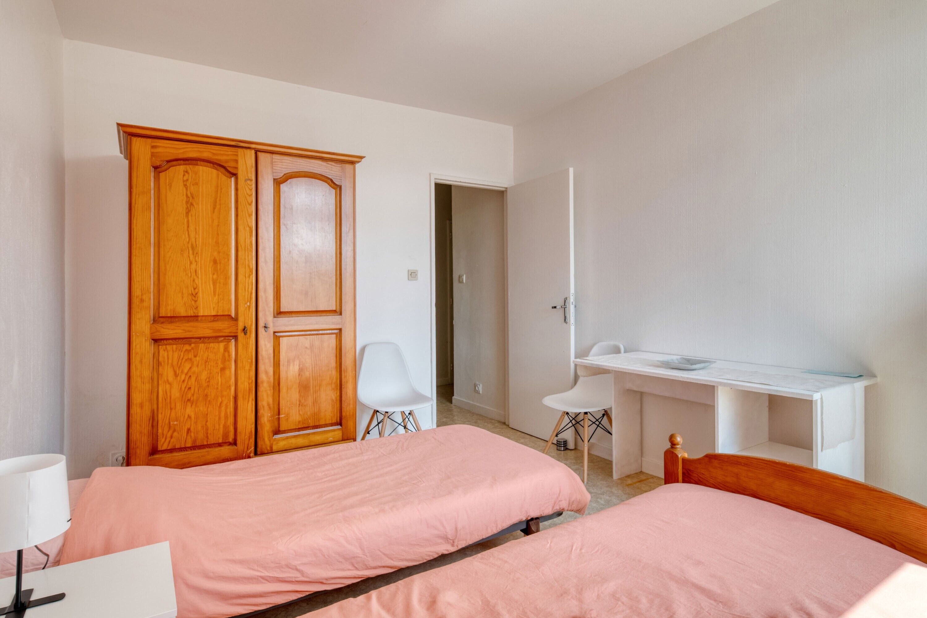 Une chambre avec deux lits, une armoire en bois, un bureau avec une chaise et un mur blanc.