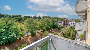 Ein Balkon mit Blick auf einen grünen Park, ein Gebäude mit roter Fassade und einem klaren, blauen Himmel.