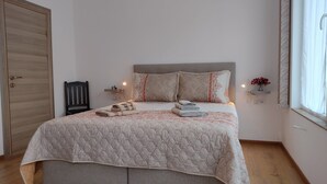 A neatly made bed with a quilted cover, two pillows, and folded towels. There is a wooden wardrobe to the left and a bedside table with a lamp and a vase of flowers to the right.