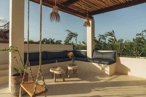 A covered outdoor seating area with a swing, two round tables, and a sofa with cushions.
