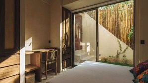 A bedroom with a bed, a desk, and a chair. There is a sliding glass door leading to a balcony with a bamboo fence.