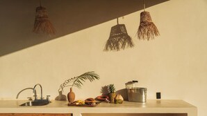 A kitchen with a sink, a plant, a fruit bowl, and hanging lights.