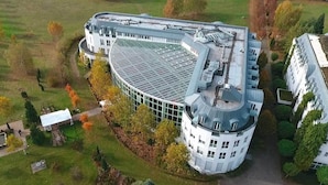 A large, circular building with a glass roof, surrounded by a well-maintained lawn and trees. - Hotel Artrium am Park (Dietzenbach)