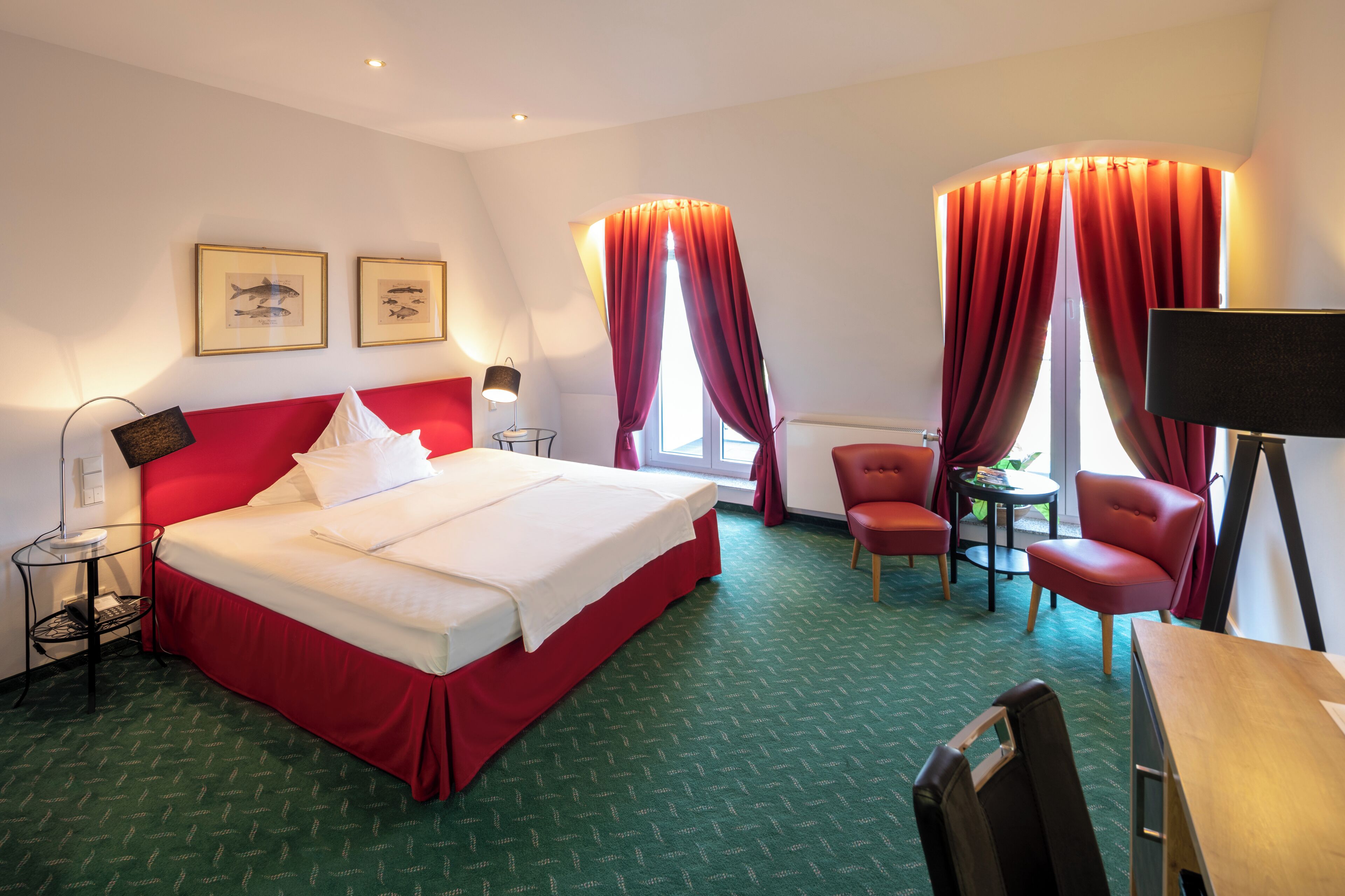 A hotel room with a large bed, a desk, two chairs, a lamp, and a window with red curtains.