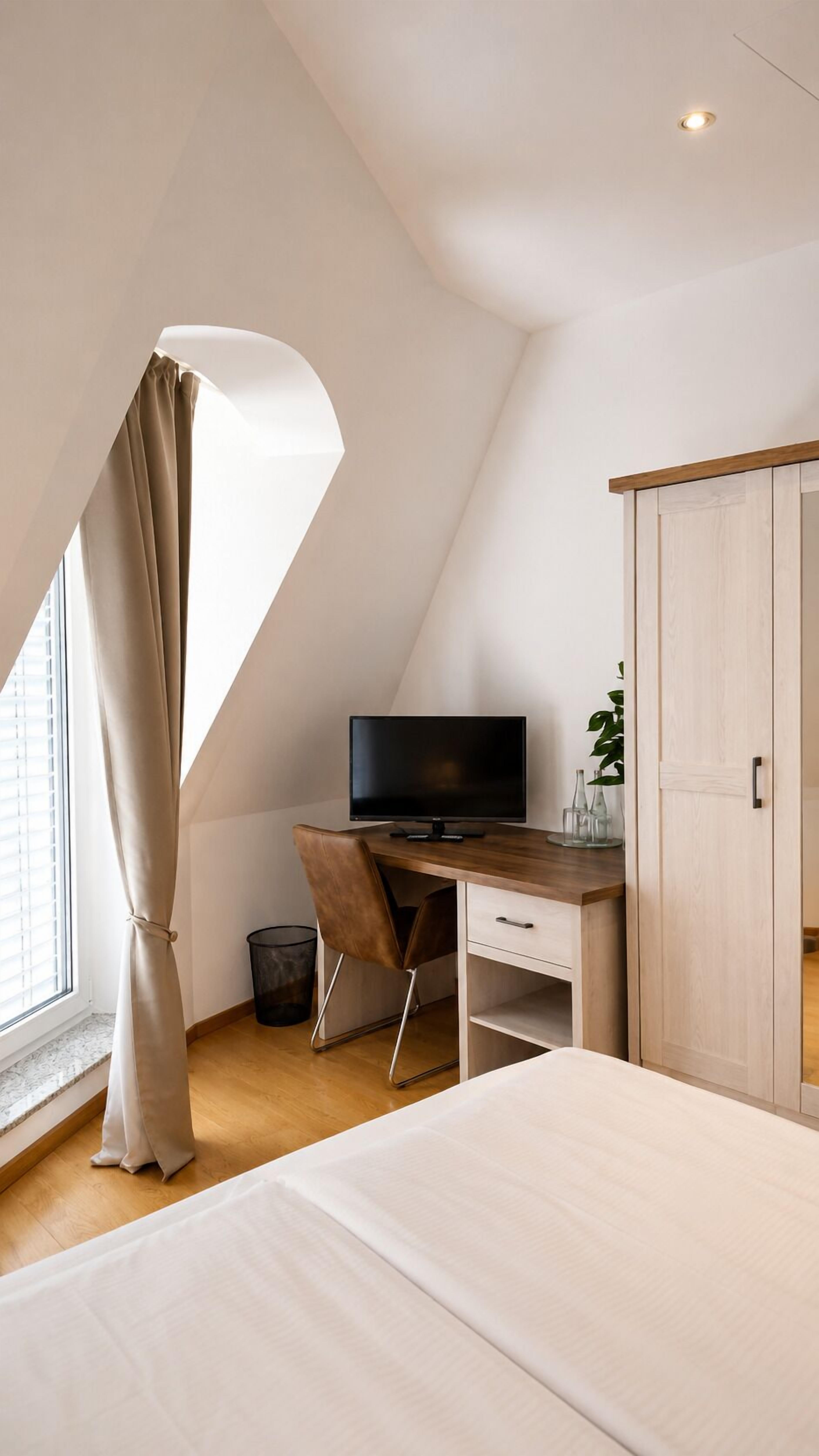 A bedroom with a bed, a desk with a TV, a wardrobe, and a window with curtains.