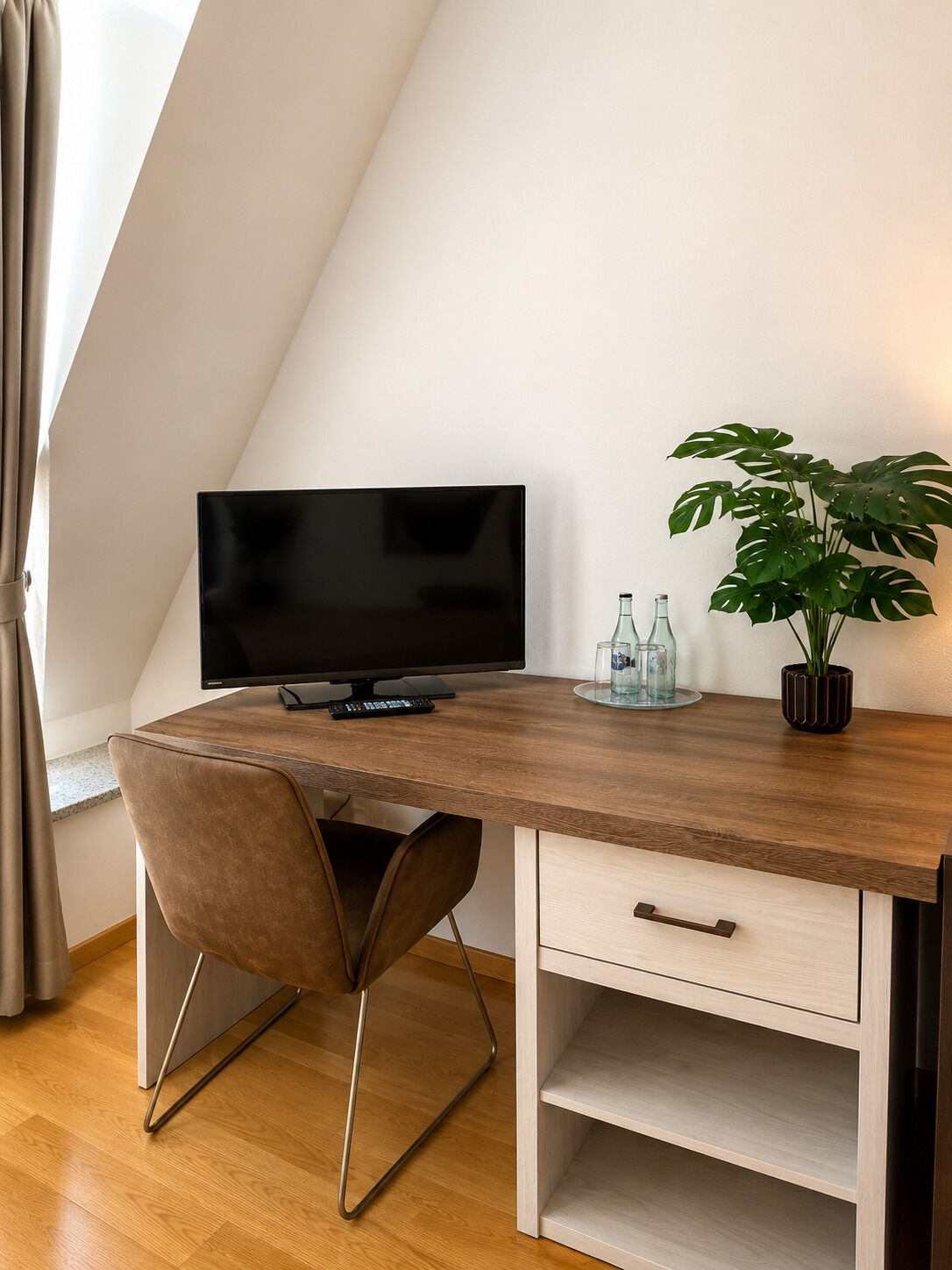 A modern, well-lit room with a wooden desk, a television, a potted plant, and a glass bottle on the desk.