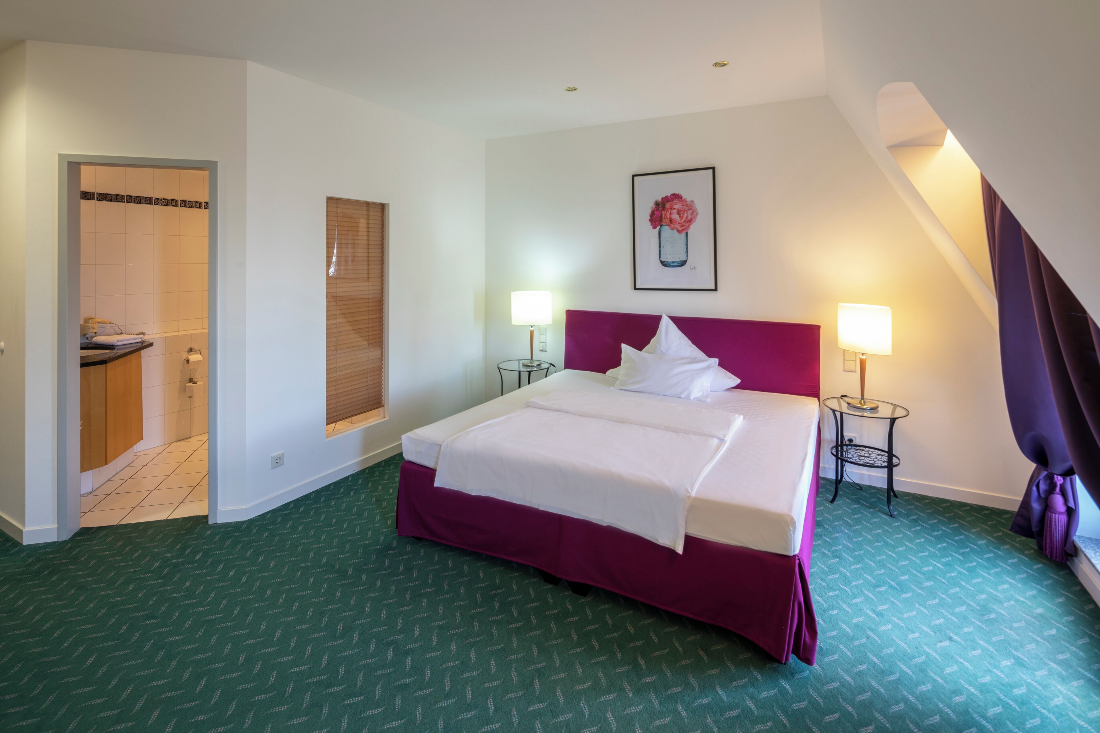 A hotel room with a large bed, two bedside tables with lamps, a framed picture on the wall, and a bathroom visible through an open door.