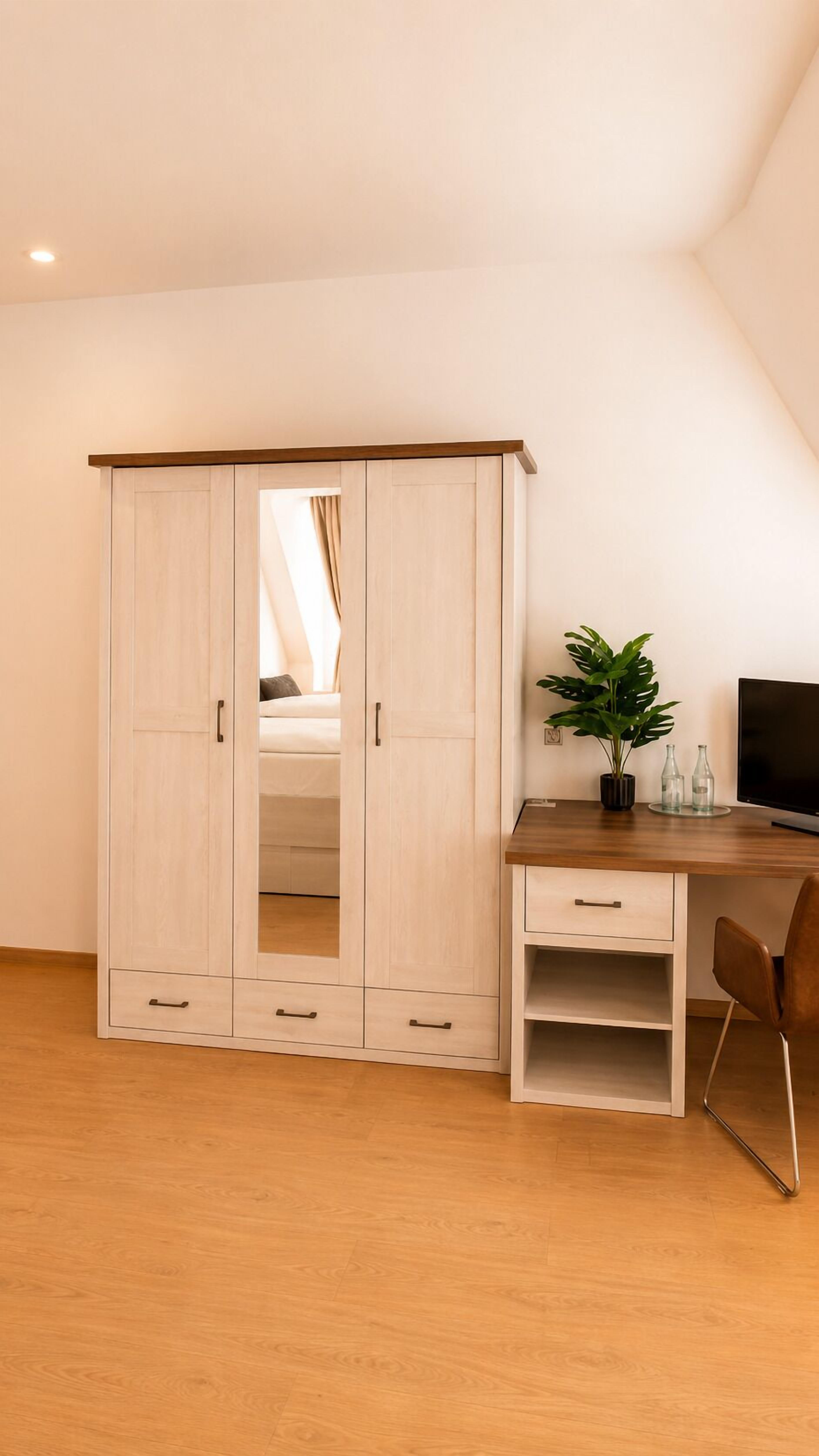 Een kamer met een houten garderobe, een bureau met stoel, een potplant en een televisie.