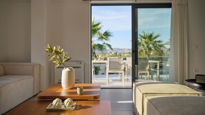 A modern hotel room with a sofa, a wooden coffee table, a vase with a plant, and a view of a balcony with outdoor furniture and palm trees.
