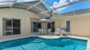A swimming pool with a glass enclosure, a covered patio area with chairs, and a door leading to a covered entryway.